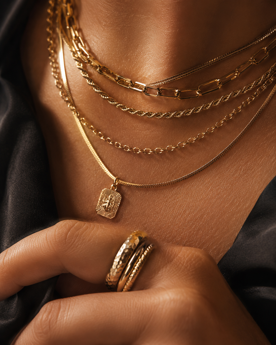Close-up of layered gold necklaces and rings worn on a model's neck and hand, with a textured gold pendant and stacked rings against a dark satin fabric background.