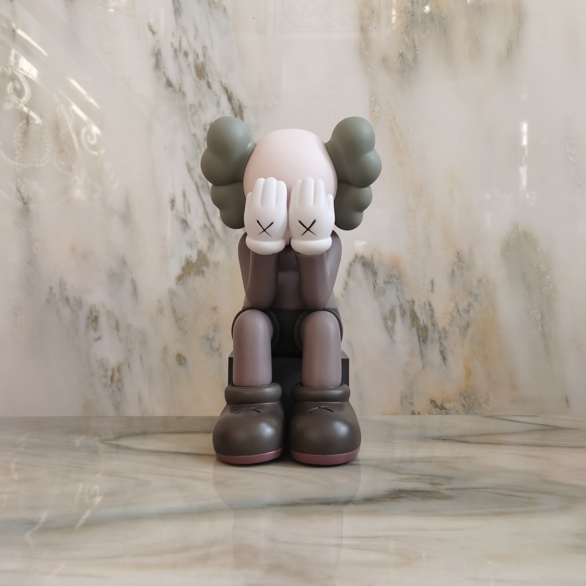 A KAWS-style collectible figure seated with its hands over its face, placed on a polished marble surface against a matching marble background.