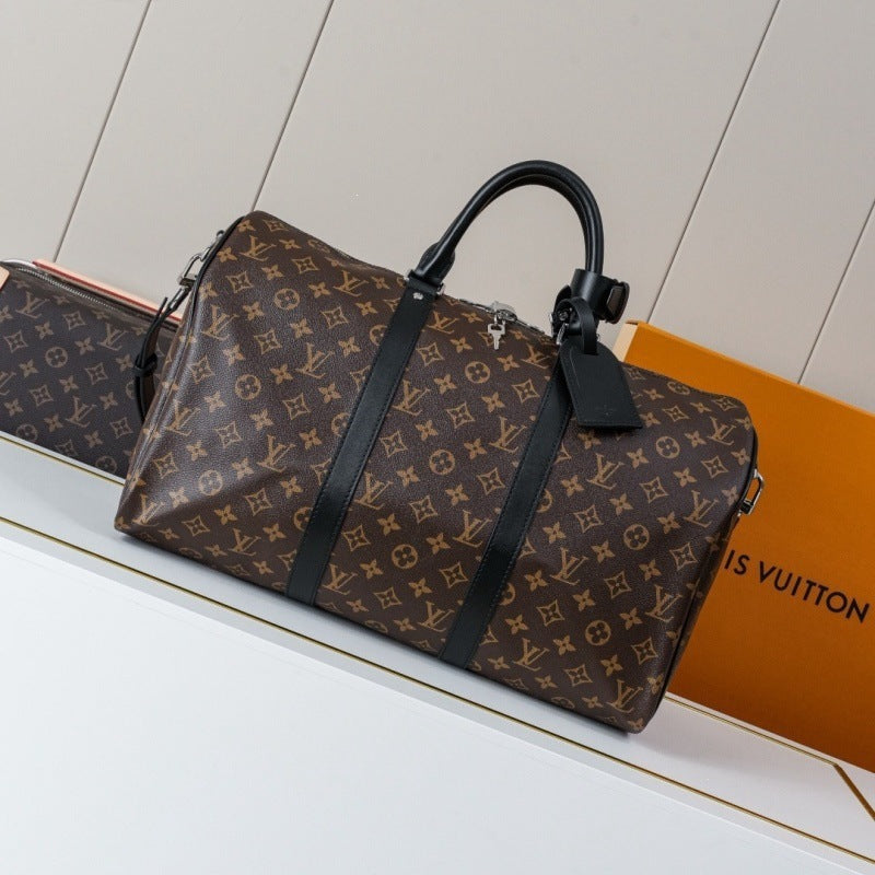 A Louis Vuitton monogram duffel bag in brown coated canvas with black leather handles and straps, displayed on a white shelf next to an orange Louis Vuitton box.