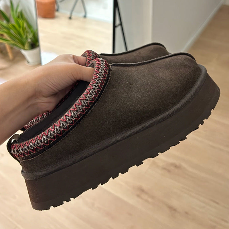 A person's hand holding a pair of dark brown suede mule slipper shoes with chunky platform soles and a red, white, and black patterned trim, set against a light wood floor background.