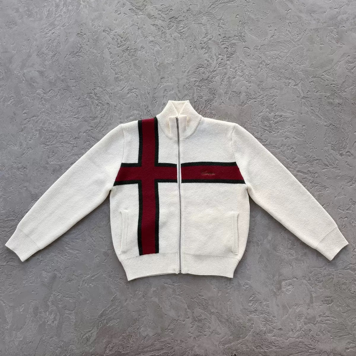A white zip-up knit cardigan sweater featuring a bold red cross design with black borders across the chest, laid flat on a textured gray background.