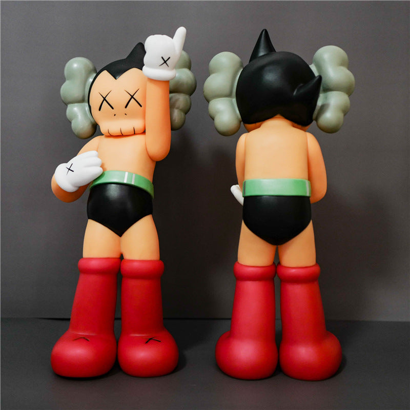 Two views of a stylized toy figure resembling Astro Boy: front view on the left and back view on the right, standing against a dark gray studio background. The figure has peach skin, black hair and shorts, red boots, a green waist band, and gray hair accessories.