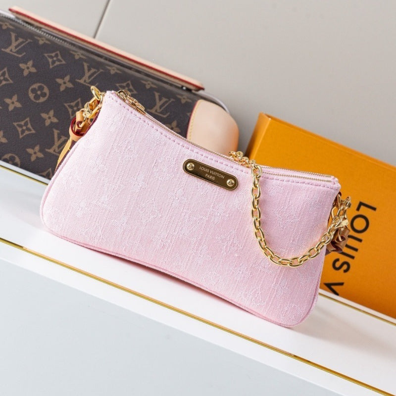 A light pink textured shoulder bag with a gold chain strap and a gold-tone metal logo plate, styled on a white surface next to a Louis Vuitton monogram pouch and an orange Louis Vuitton box.