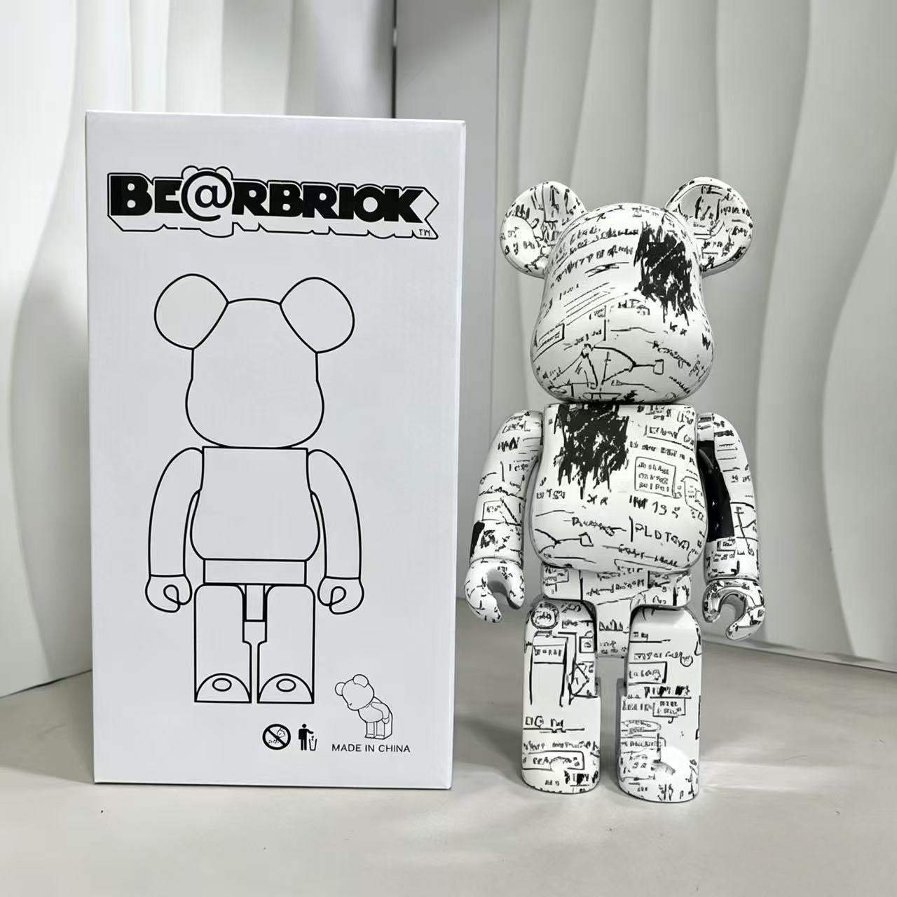 A Bearbrick collectible figure with a white body covered in black graffiti-style text and scribbles, standing next to its white retail box featuring a black line drawing of the bear figure and the BE@RBRIOK logo.