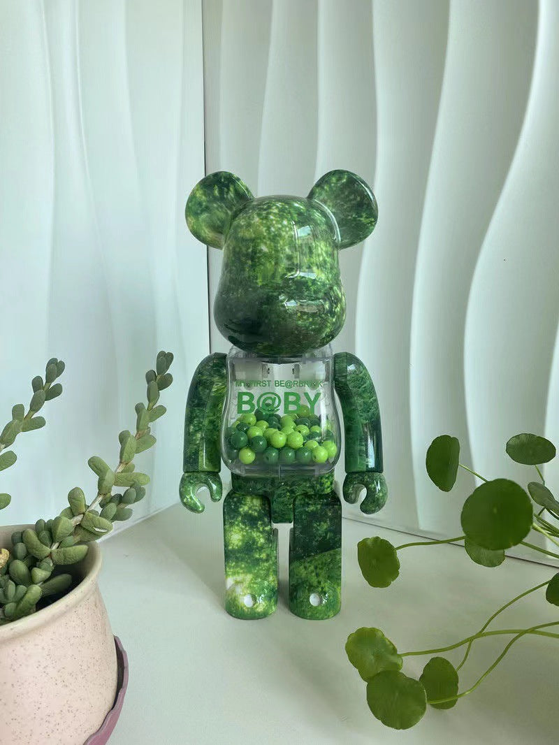 A green marbled bear-shaped collectible figure with a transparent chest panel filled with small green beads, standing on a white surface with potted plants and white wavy curtains in the background.