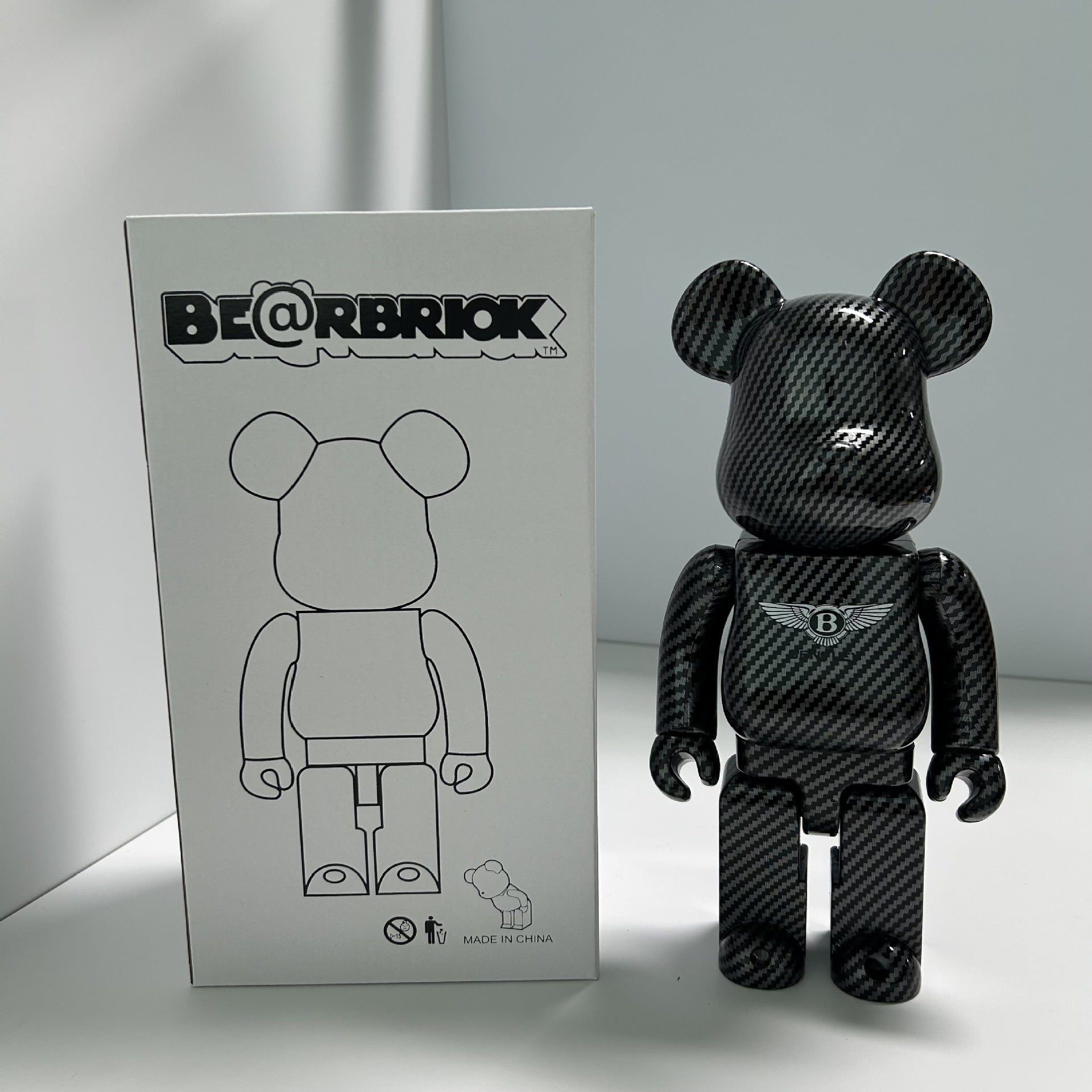 A black carbon-fiber-patterned Bearbrick toy figure stands next to its white packaging box, which features a line drawing of the bear figure and the BE@RBRIKK brand name.