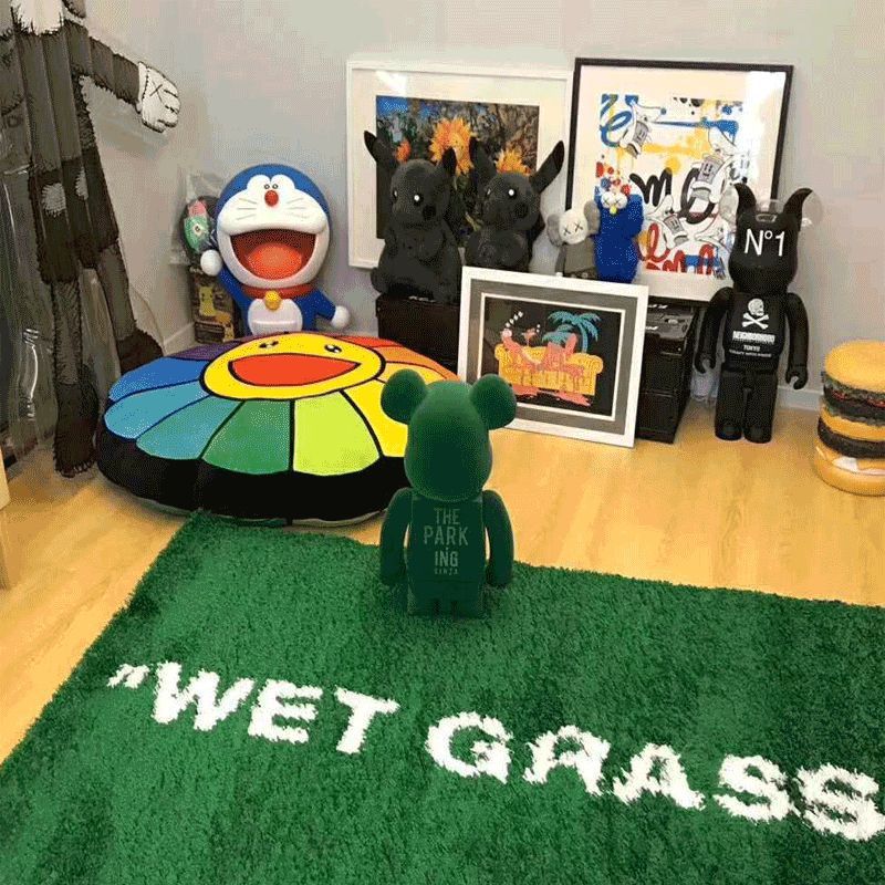 A green shag rug with white text reading 'WET GRASS' is placed on a light wood floor in a styled room. A small green bear-shaped figure stands on the rug.