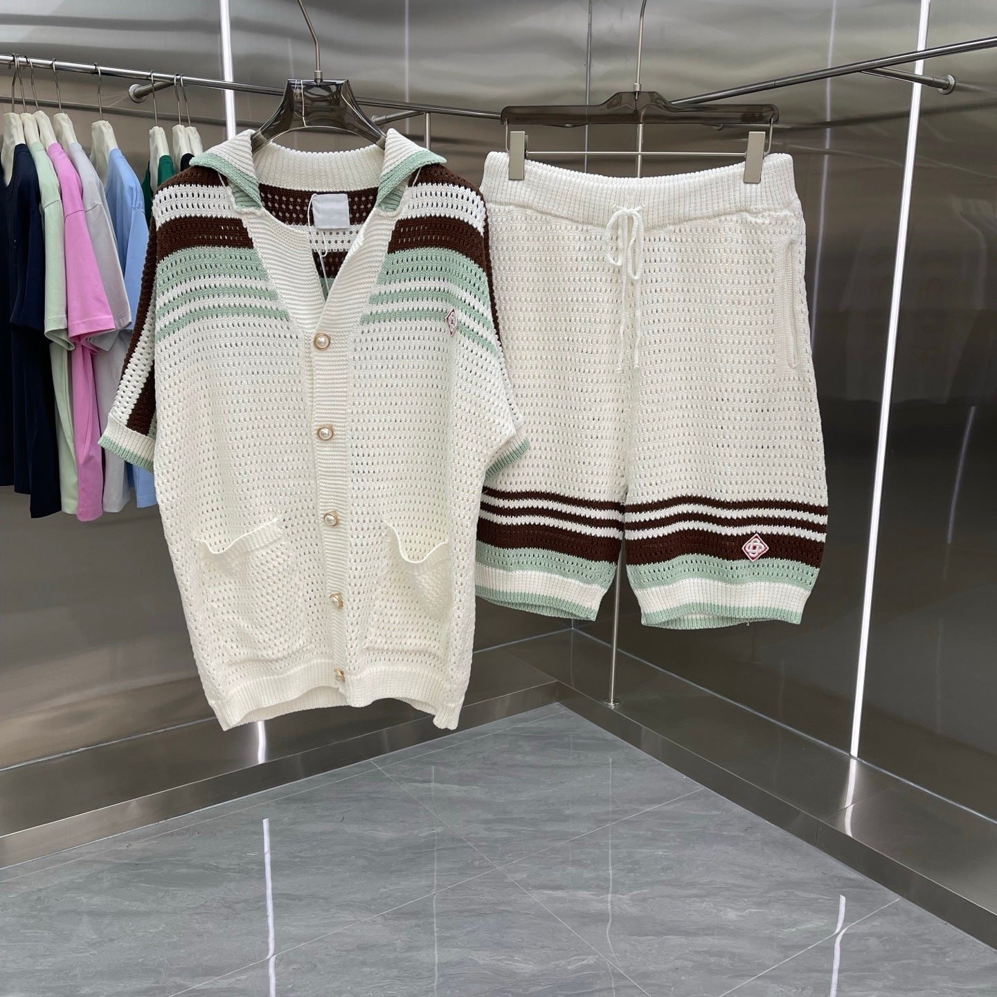 A knit two-piece set consisting of a short-sleeve button-front cardigan and matching shorts, hanging on a rack in a store. The set is primarily off-white/cream with horizontal stripes in brown, sage green, and light blue.