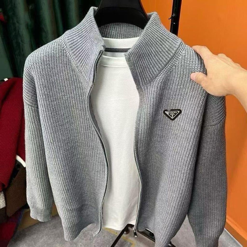 A light gray ribbed knit zip-up jacket with a stand collar and a small black triangle logo patch on the chest, displayed on a hanger over a white t-shirt. A hand is holding the sleeve of the jacket.
