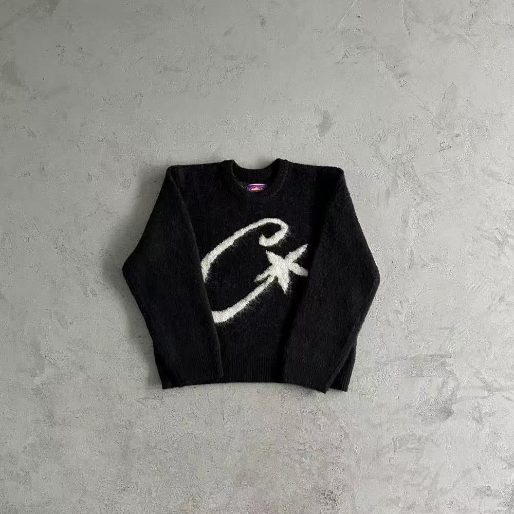 A black knit crewneck sweater with a large white abstract graphic on the front, laid flat on a textured gray concrete surface.