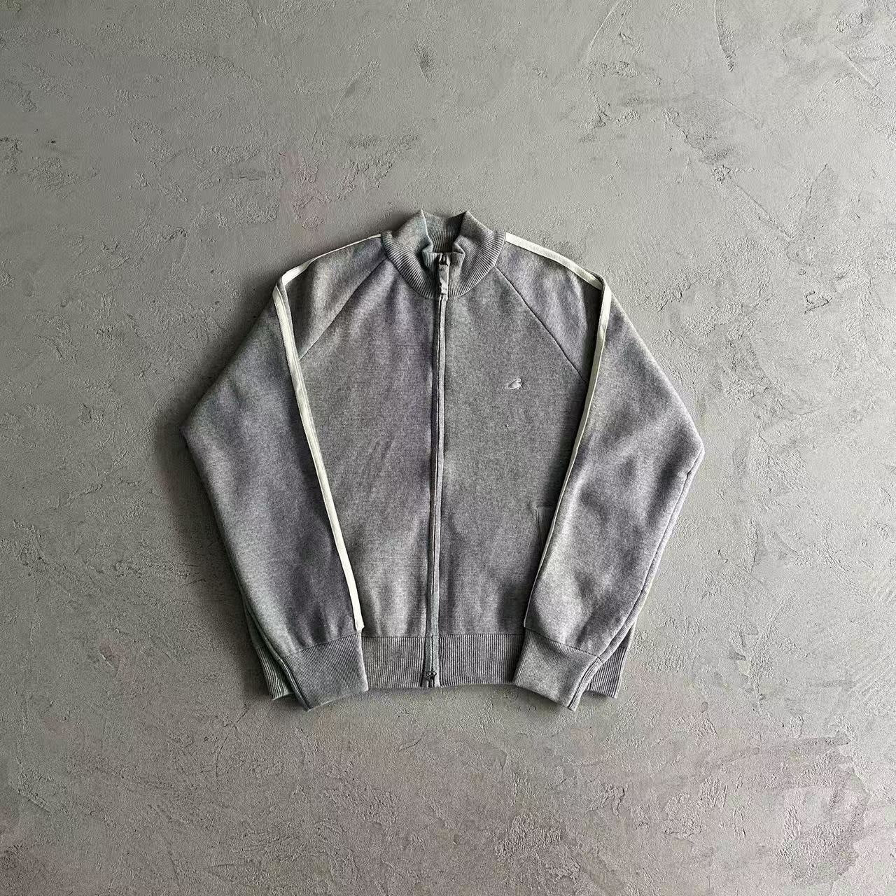 A gray zip-up track jacket with white stripes on the shoulders, laid flat on a textured gray concrete surface.