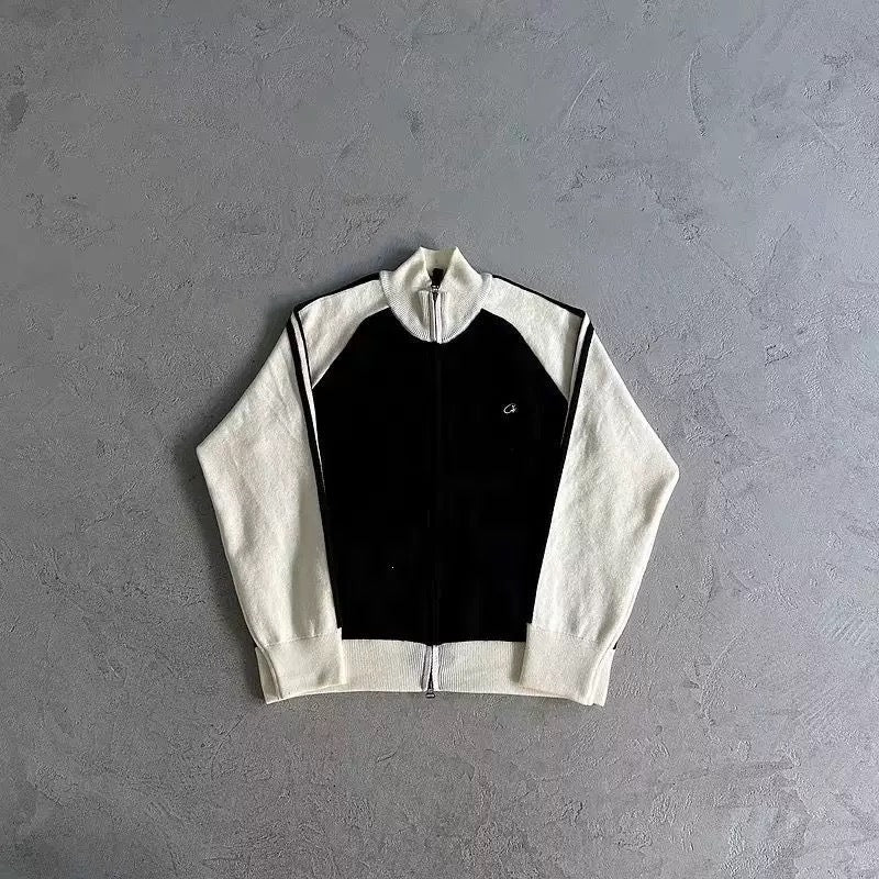 A colorblock zip-up track jacket laid flat on a textured gray concrete background. The jacket features an off-white/cream body and sleeves with a solid black central panel, accented by thin black stripes on the shoulders and a small white embroidered logo on the left chest.