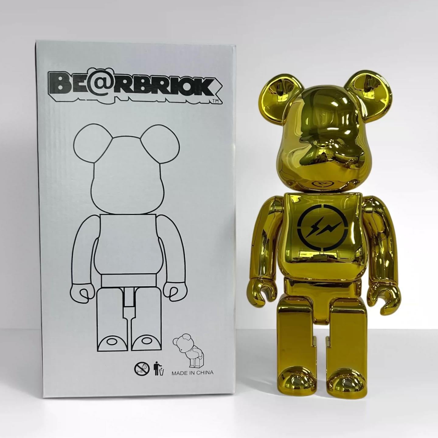 A shiny metallic gold Bearbrick toy stands next to its original white packaging box, which features the 'BE@RBRIOK' logo and a black outline drawing of the bear figure.