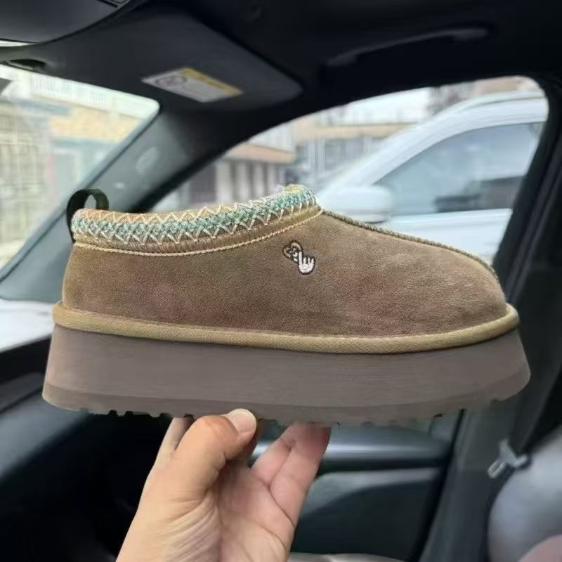 A person holds up a brown suede slipper shoe inside a car, showing the side profile with a thick brown sole, a light beige trim, and a small embroidered logo.