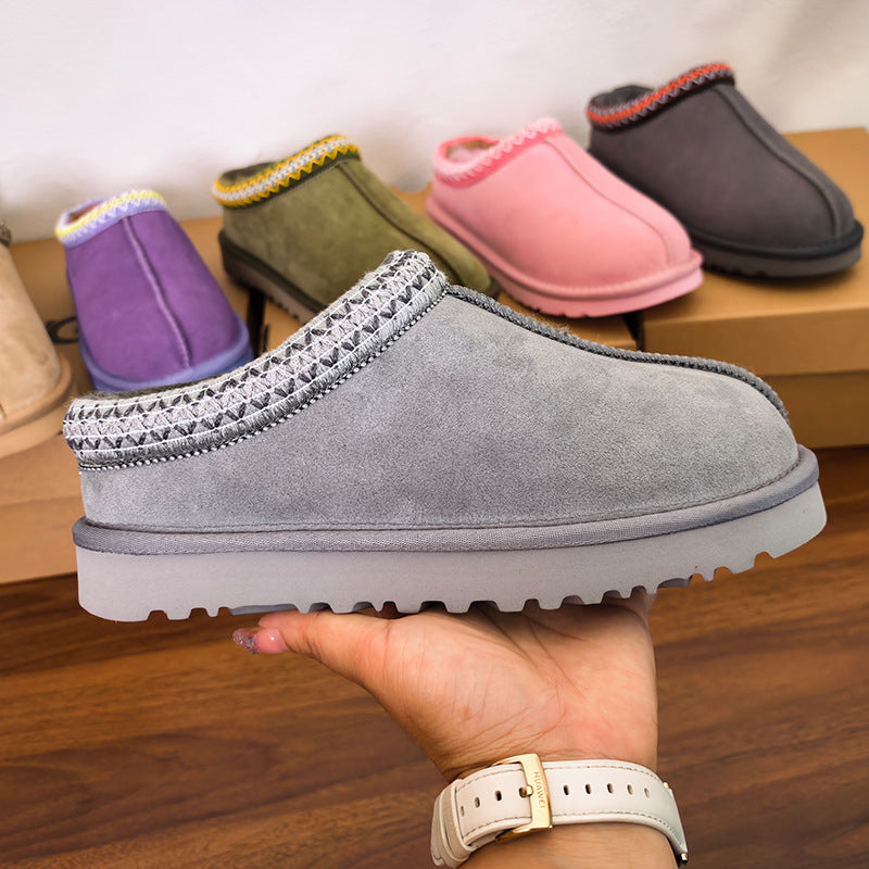 A hand holds a gray suede slip-on mule bootie with a thick sole and braided trim, with several other color options displayed in the background on a wooden surface.