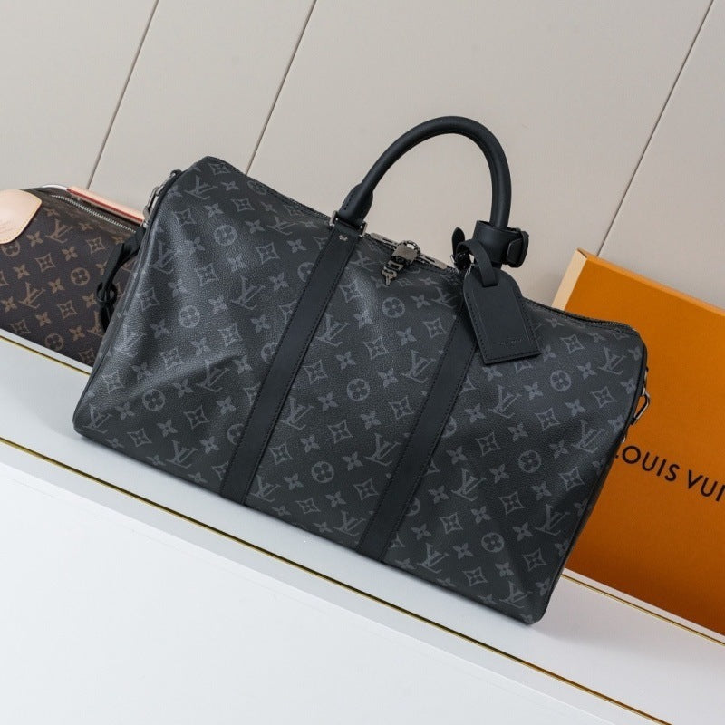 A Louis Vuitton monogram black duffel bag with black leather handles and trim, displayed on a white shelf next to an orange Louis Vuitton box.