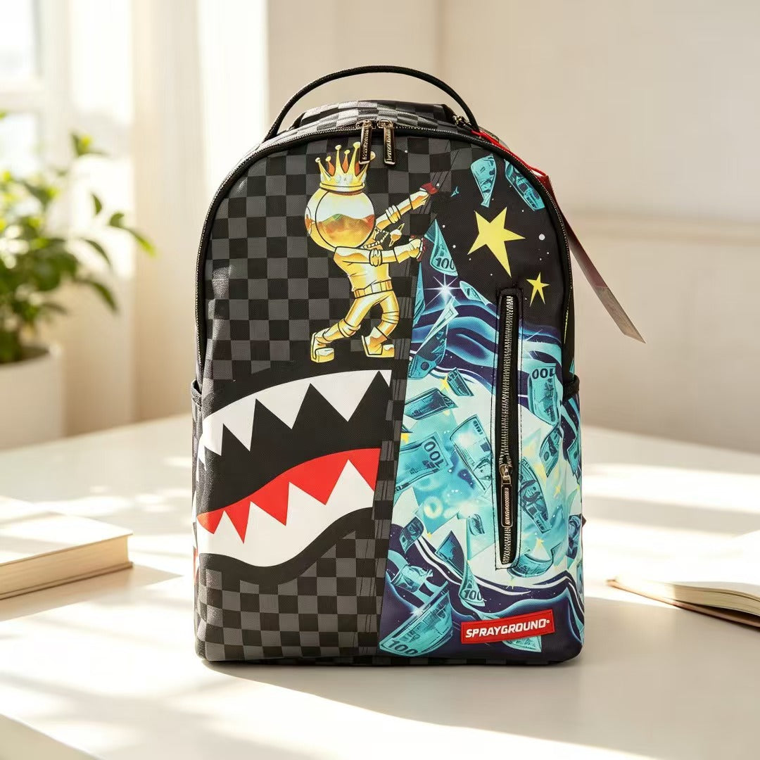 A Sprayground backpack with a split design: one half features a black and gray checkerboard pattern with a gold astronaut graphic, and the other half features a blue and black dollar-bill pattern. The bottom has a red Sprayground logo patch, and the bag is shown on a table in a bright room.