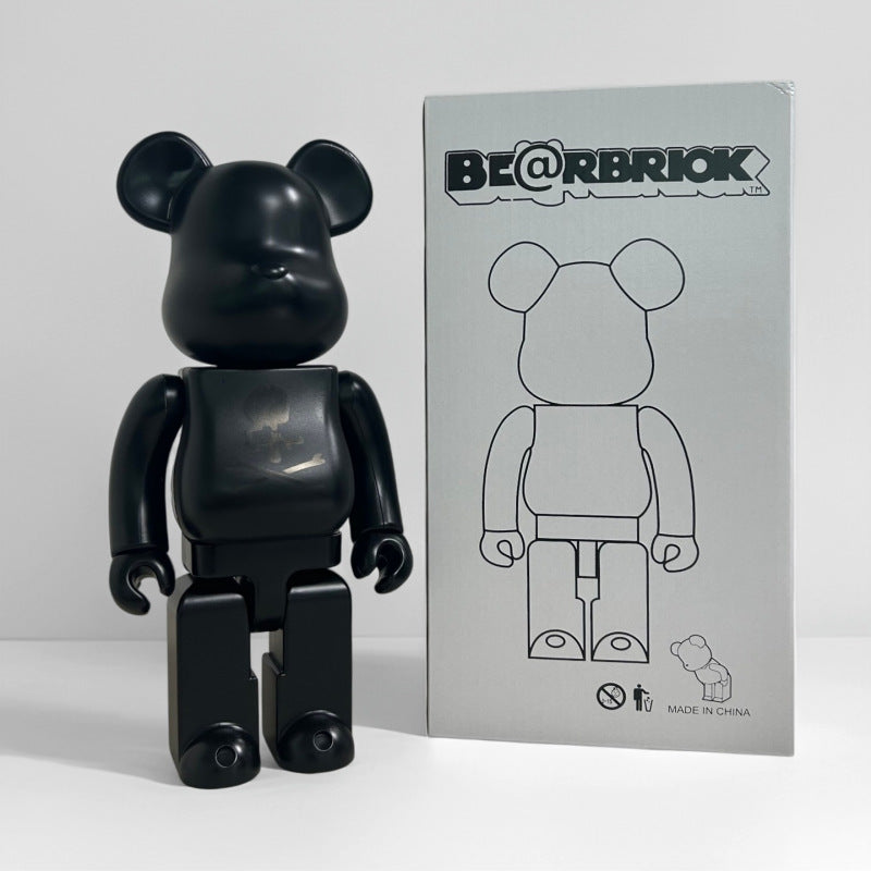 A matte black Bearbrick collectible figure standing next to its light gray packaging box, which features the BE@RBRIOK logo and a line drawing of the bear figure.