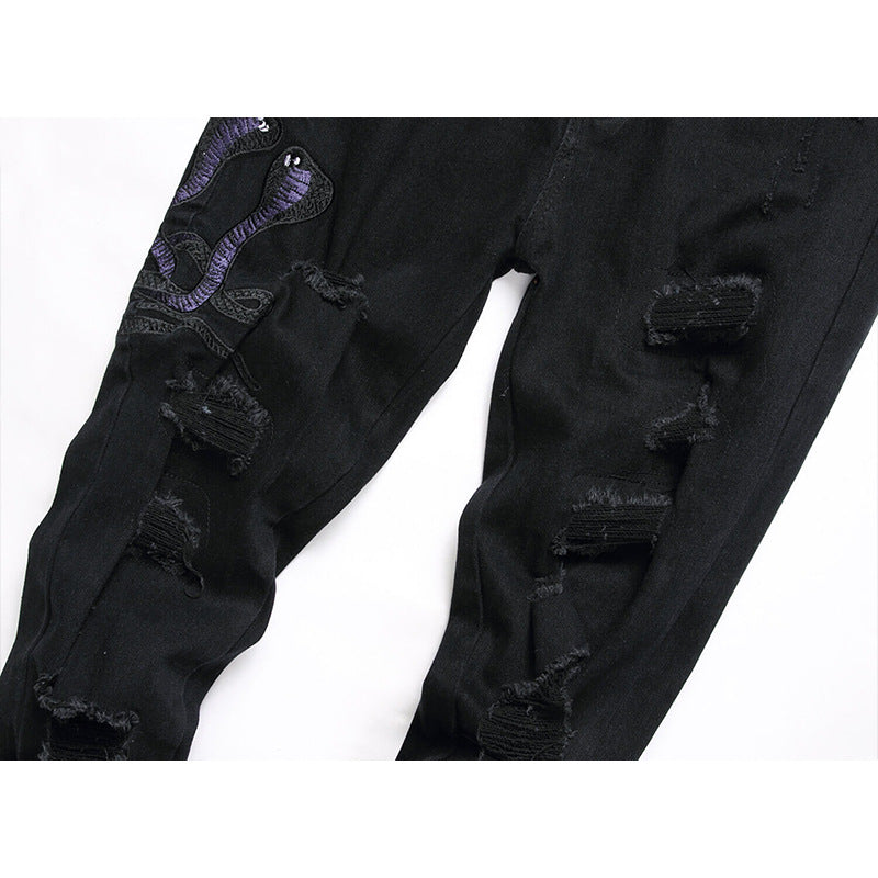 Black distressed denim jeans with ripped details and a purple embroidered snake design on the upper leg, shown against a plain white background.