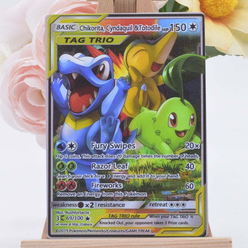 A Pokémon TCG Chikorita card displayed upright on a small wooden stand, with pink flowers and a yellow flower blurred in the background.