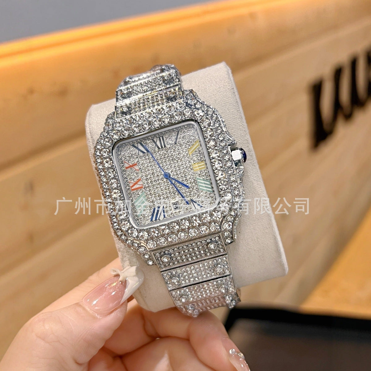 A square-shaped watch covered in sparkling rhinestones, held by a hand against a warm wooden background. The watch has a silver-toned crystal-encrusted bezel, a matching crystal-embellished link band, and a light-colored dial with multicolor Roman numerals.