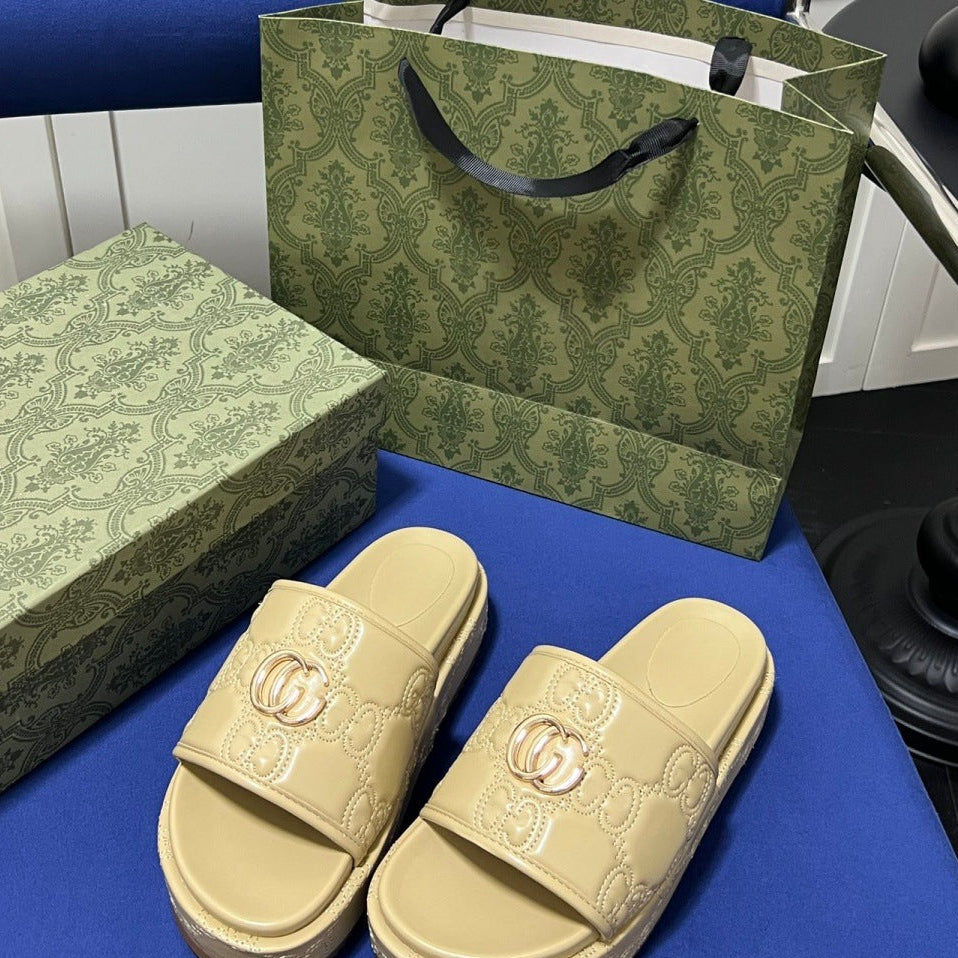 A pair of beige platform slide sandals with embossed monogram pattern and a gold-tone interlocking G logo, displayed next to a green patterned Gucci box and matching shopping bag on a blue surface.