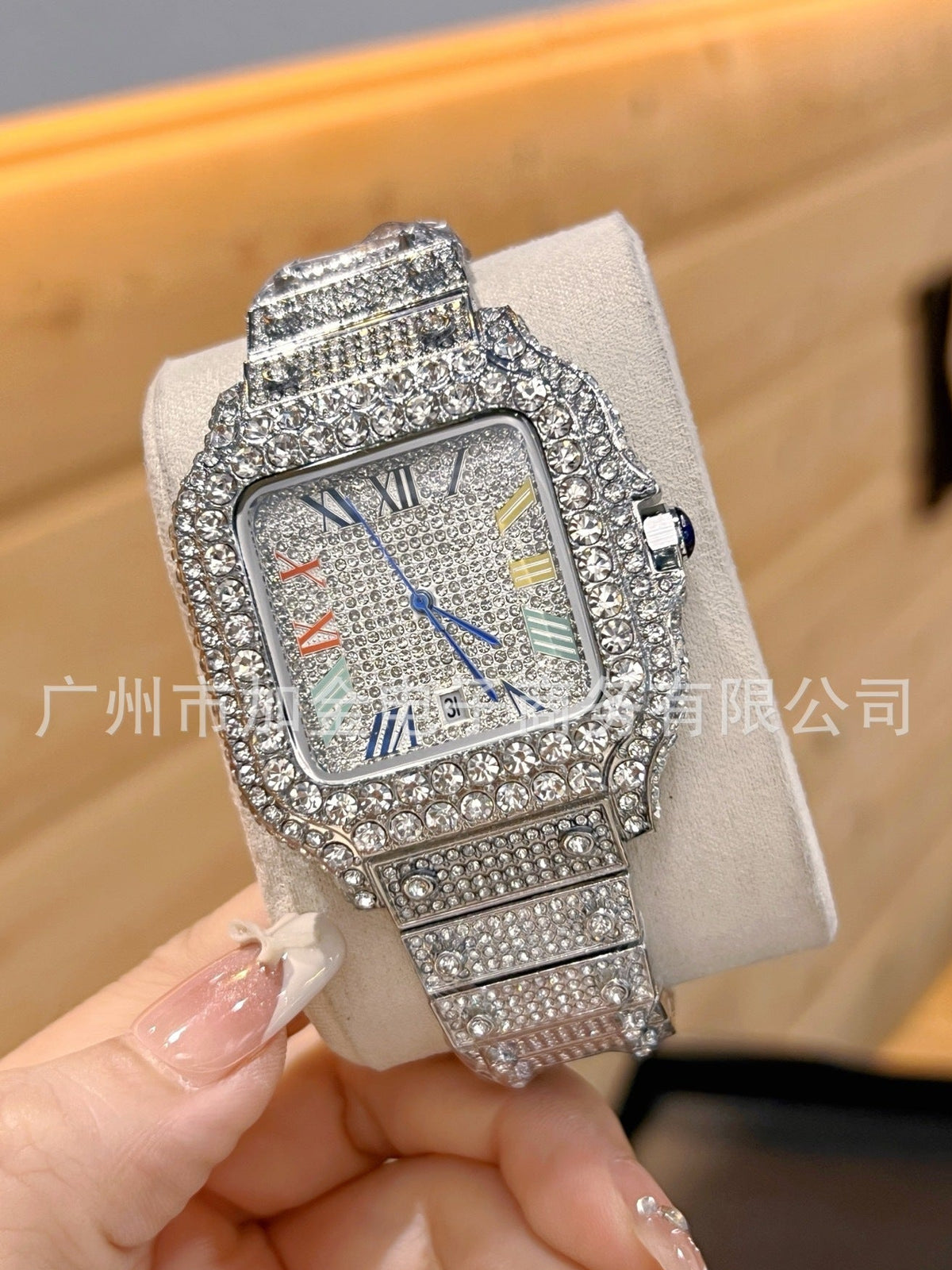 A heavily rhinestone-embellished silver-tone watch with a square bezel and matching link bracelet, displayed on a light beige jewelry stand and held by a hand. The watch face has a textured silver dial with multicolored Roman numerals and blue hands.