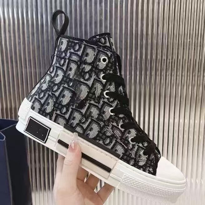 A high-top sneaker with a black and white repeating monogram pattern, held by a hand against a metallic ribbed background.