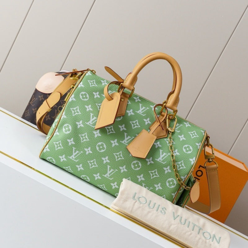 A green Louis Vuitton monogram Speedy handbag with tan leather handles and trim, displayed on a white shelf with a Louis Vuitton dust bag and orange box in the background.