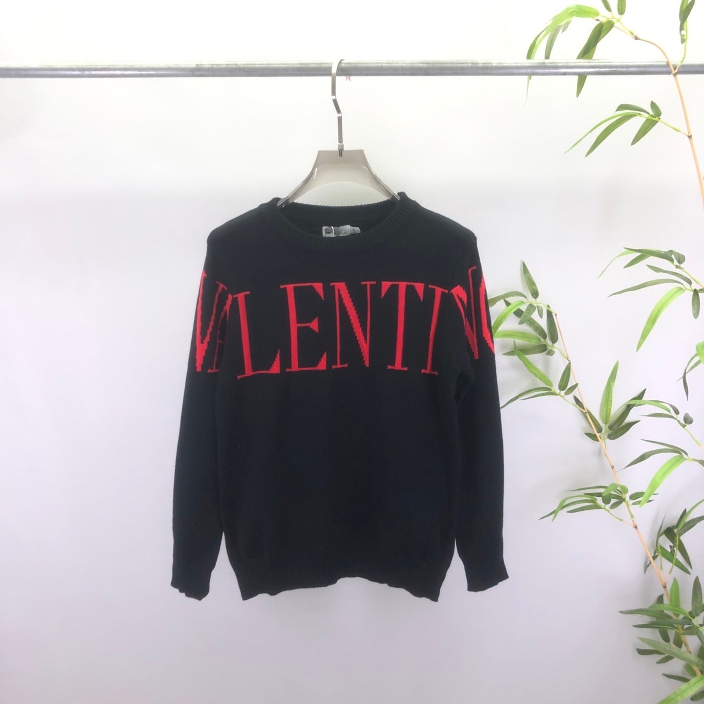 A black long-sleeve crewneck sweatshirt with 'VALENTINO' printed across the chest in red, hanging on a hanger against a white wall with a green plant on the right.