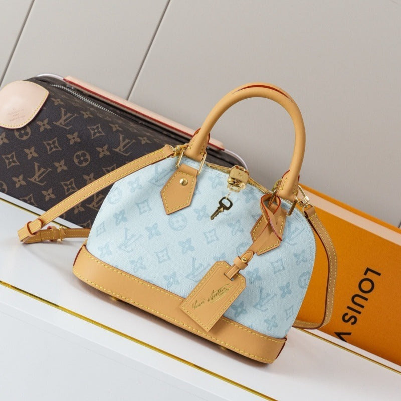 A Louis Vuitton Speedy handbag in light blue monogram canvas with tan leather trim and handles, displayed on a white surface next to an orange Louis Vuitton box and a brown monogram pouch.