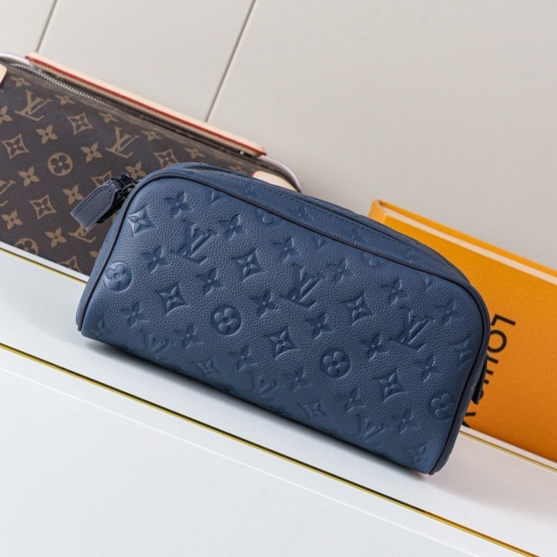 A Louis Vuitton monogram canvas pouch in dark brown with a pink zipper, displayed next to a navy blue leather zippered pouch featuring an embossed monogram pattern, with an orange Louis Vuitton box in the background.