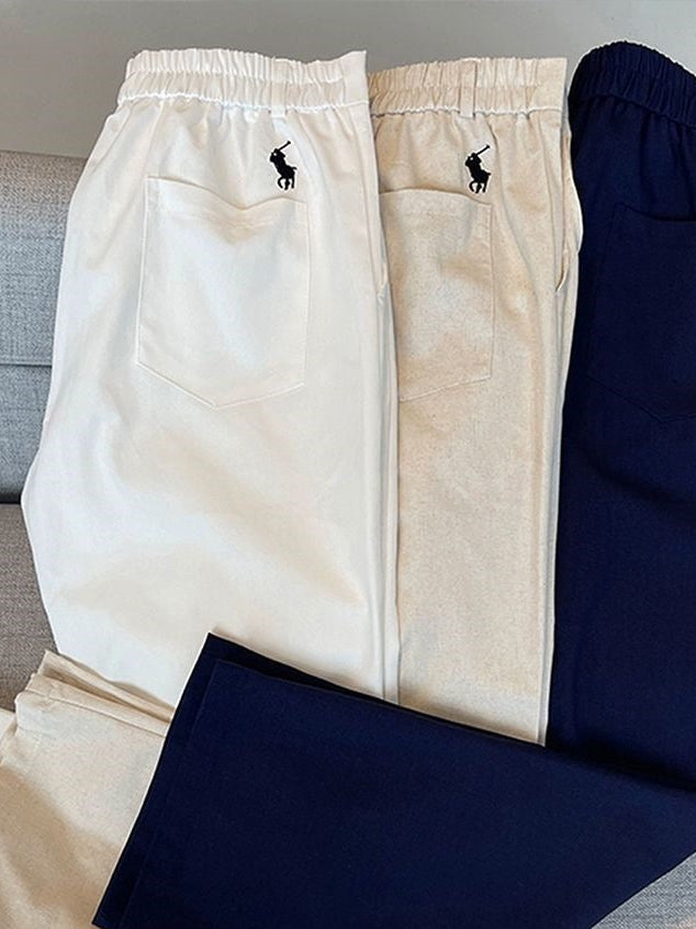 Three pairs of Polo Ralph Lauren elastic-waist trousers in cream, light beige, and navy blue, folded and arranged on a gray sofa. The Polo Ralph Lauren pony logo is visible on the back pockets of each pair.