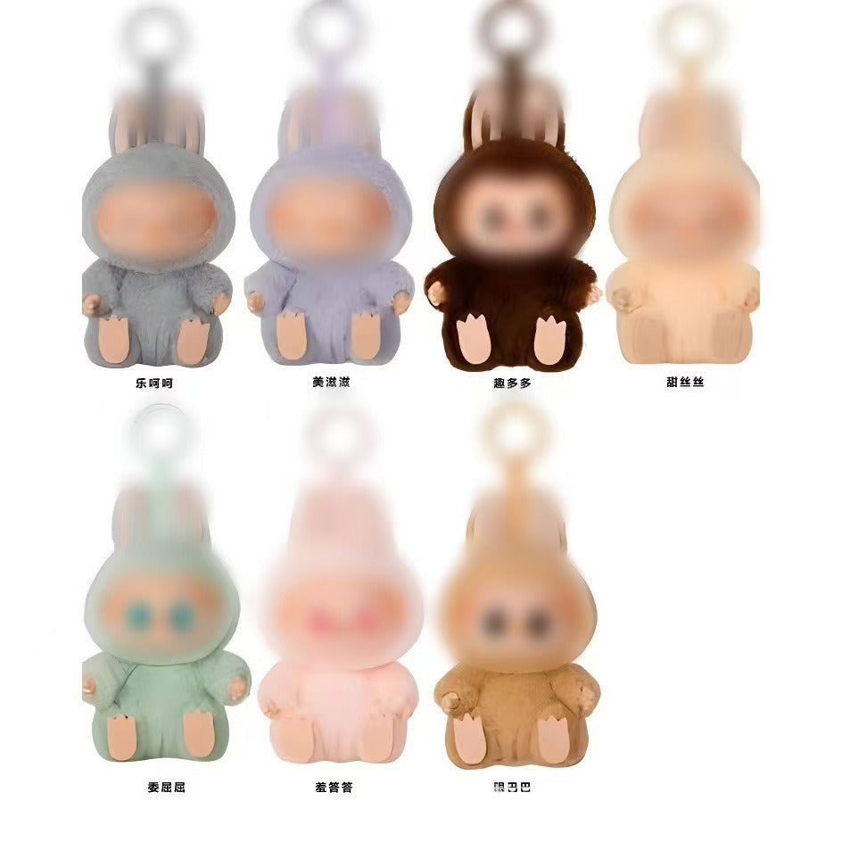 Six small plush character keychain toys arranged in two rows on a white background, each with a different soft color: gray, lavender, dark brown, cream, mint green, pale pink, and tan. Each toy has a fuzzy hooded body, rounded face, small hands, and a loop at the top for a keychain.