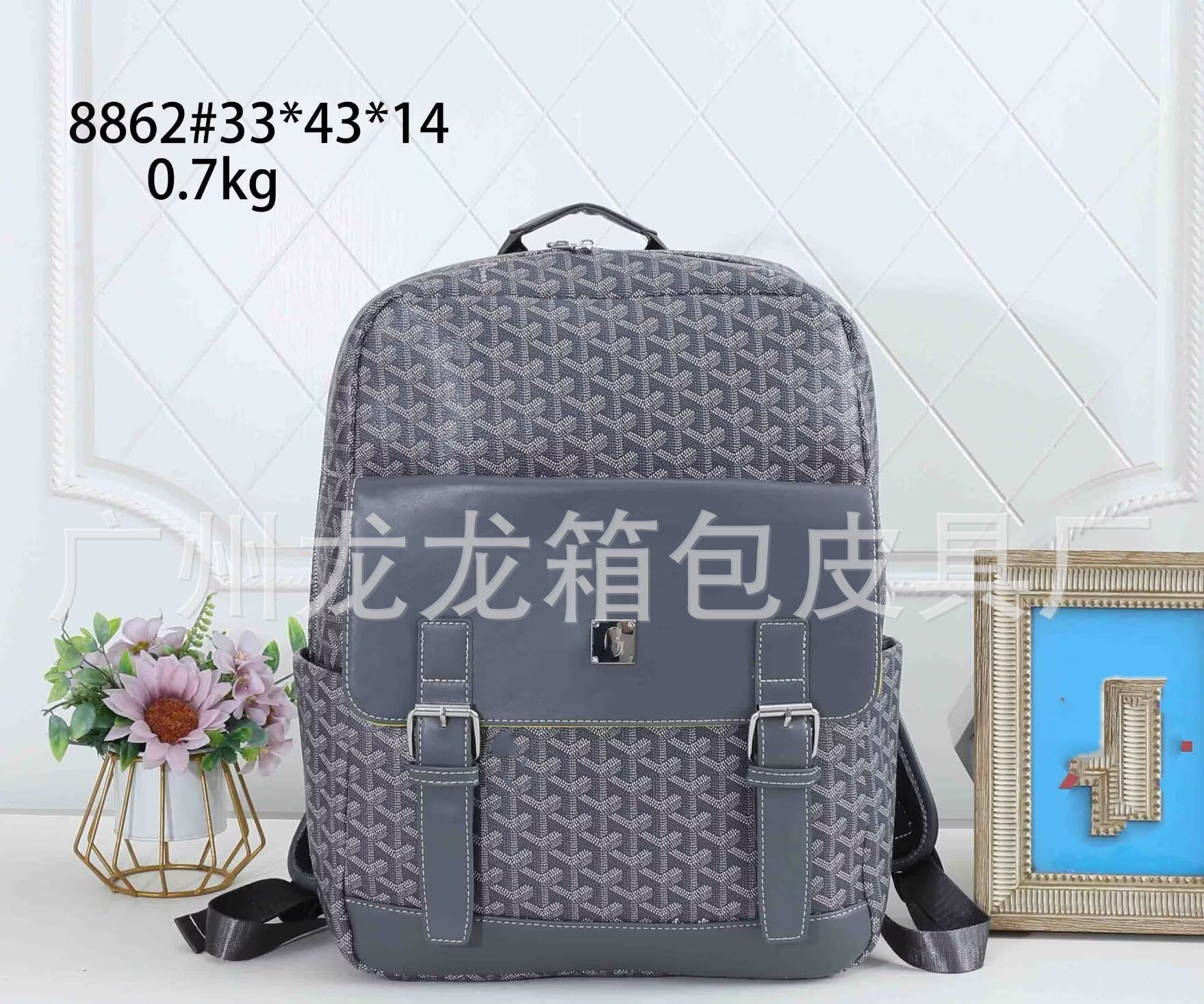 A gray patterned backpack with a dark gray front flap and buckle straps, displayed on a white surface with decorative props including a small flower vase and a framed picture. Text overlays show product code and dimensions.