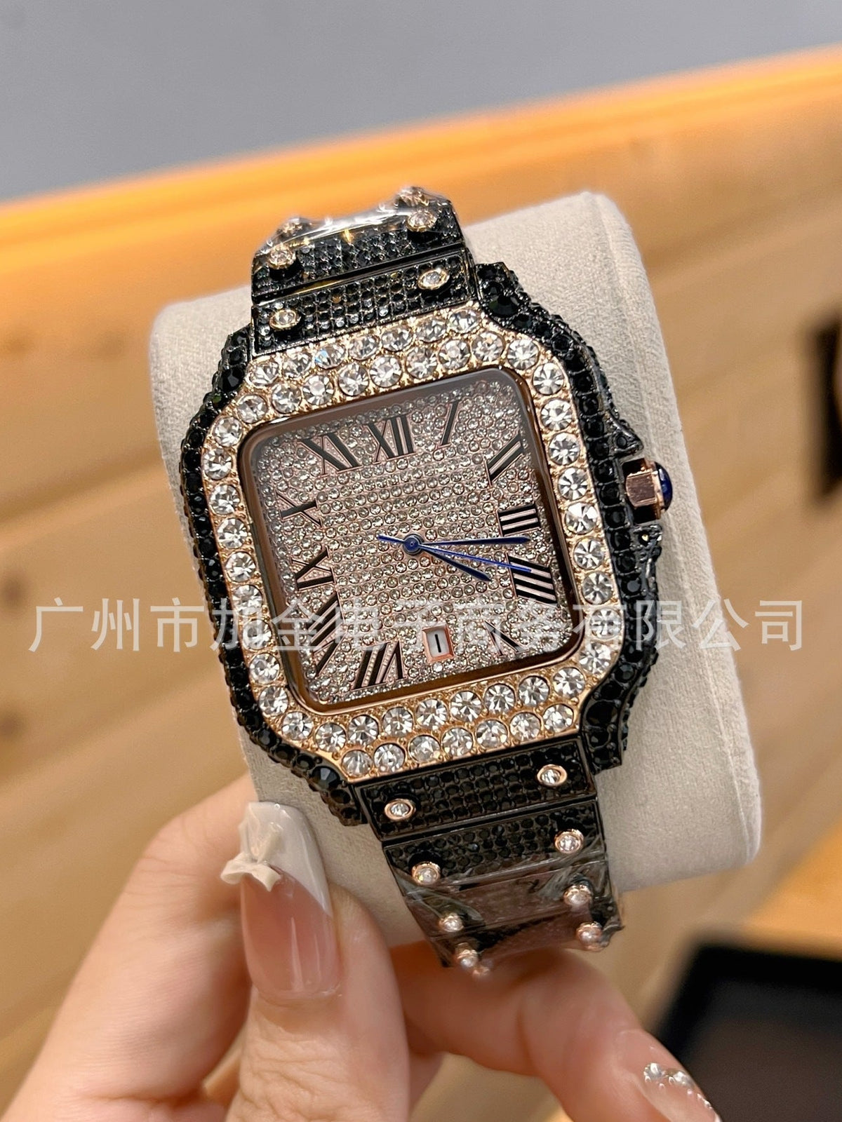 A luxury-style rectangular watch with a textured silver dial, a black rhinestone-embellished bezel and strap, and blue hands, held in a hand against a warm wood background.