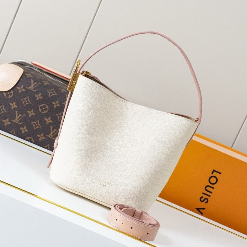 A cream-colored pebbled leather Louis Vuitton bucket bag with a light pink top handle and trim, displayed next to its brown monogram pouch, an orange Louis Vuitton box, and a matching pink strap on a white surface.