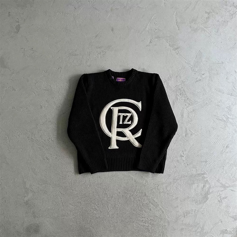A black ribbed crewneck sweater with a large white 'CR' monogram logo featuring the letters 'TZ' inside, laid flat on a textured gray concrete surface.