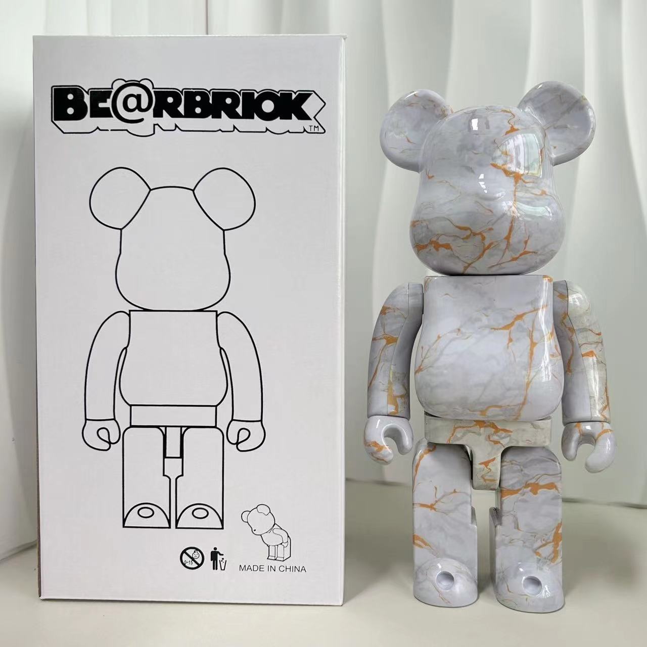 A Bearbrick collectible figure with a white and gray marble pattern and orange veins, standing next to its white packaging box featuring a black line drawing of the figure.