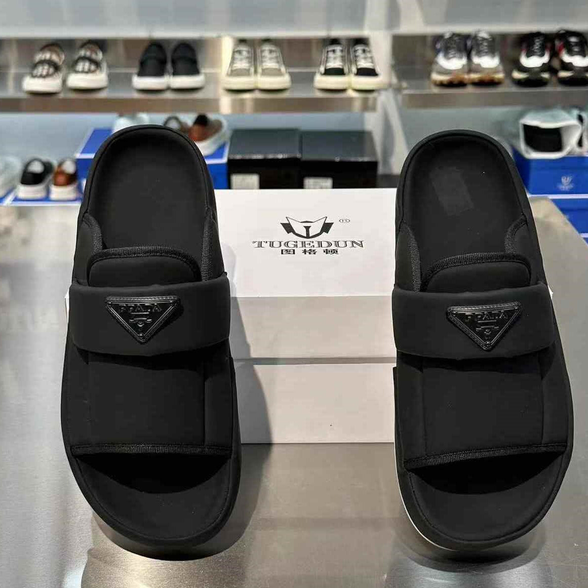 A pair of black slide sandals displayed side by side on a metallic counter, with a white shoebox in the background. The sandals feature a wide strap with a triangular logo emblem, and the background shows a retail shoe display.