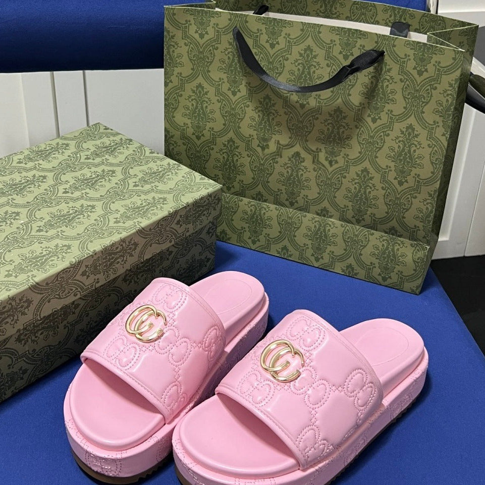 Pink platform slide sandals with embossed monogram pattern and gold double-G hardware, shown with a matching green patterned shoe box and shopping bag on a blue surface.