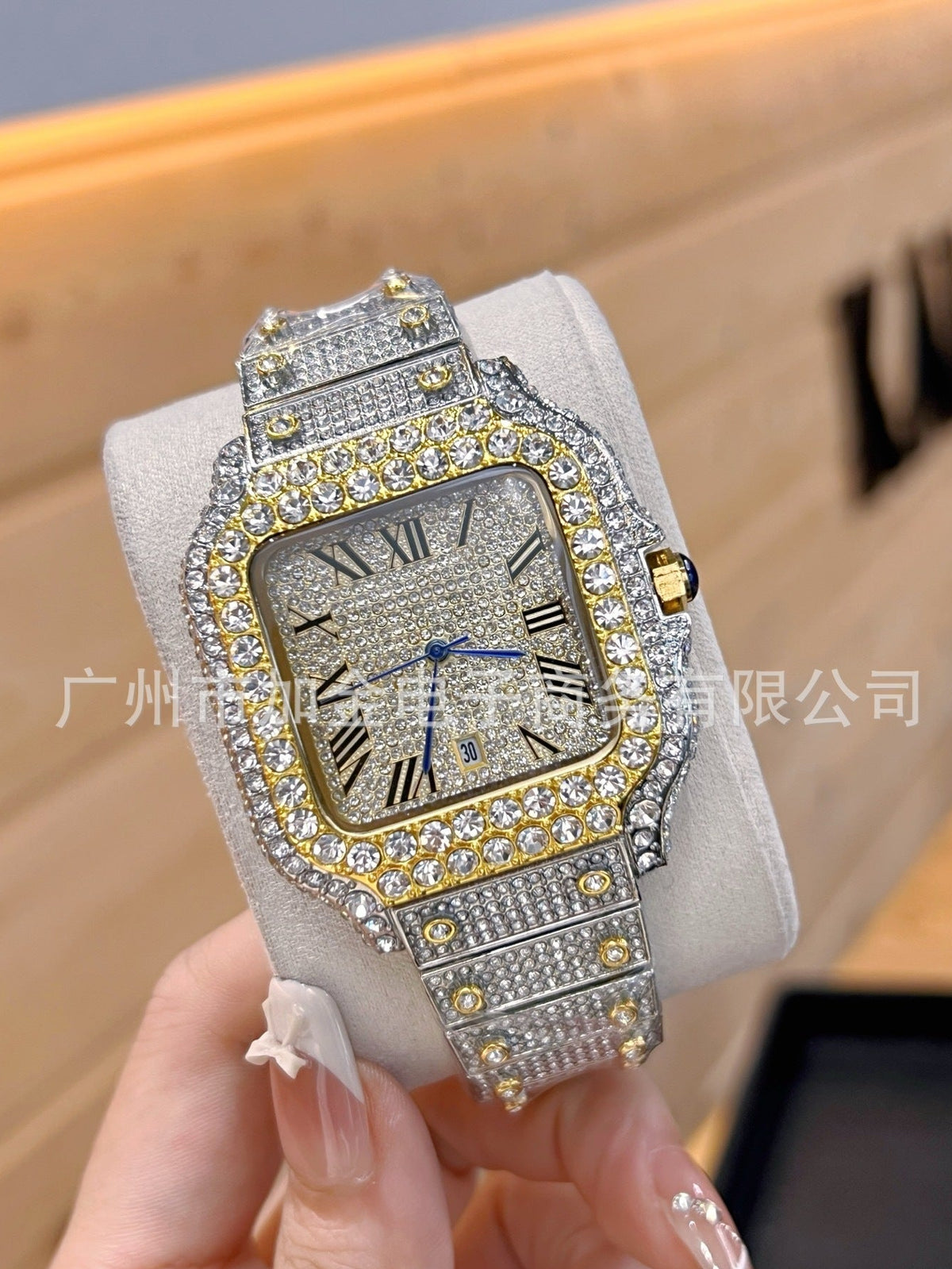 A heavily embellished rectangular watch with a silver-toned case and bracelet covered in clear stones, gold-toned accents around the bezel and crown, and a gray dial with black Roman numerals. The watch is displayed on a light gray cushion held by a hand.