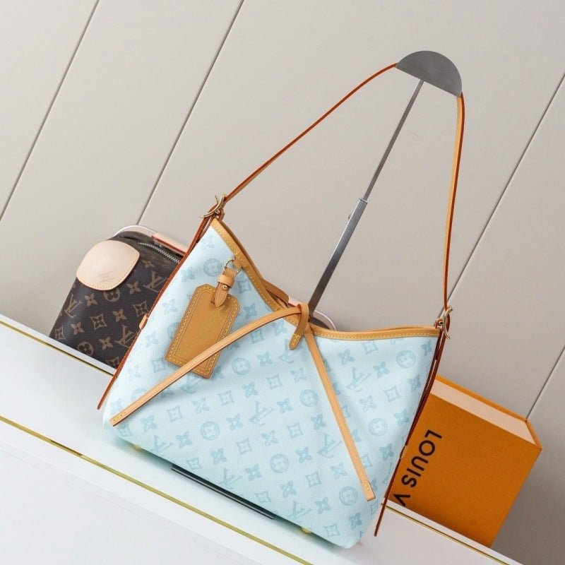 A light blue Louis Vuitton Neverfull tote bag with tan leather trim and monogram pattern, displayed on a white shelf next to a Louis Vuitton box and another monogram bag.