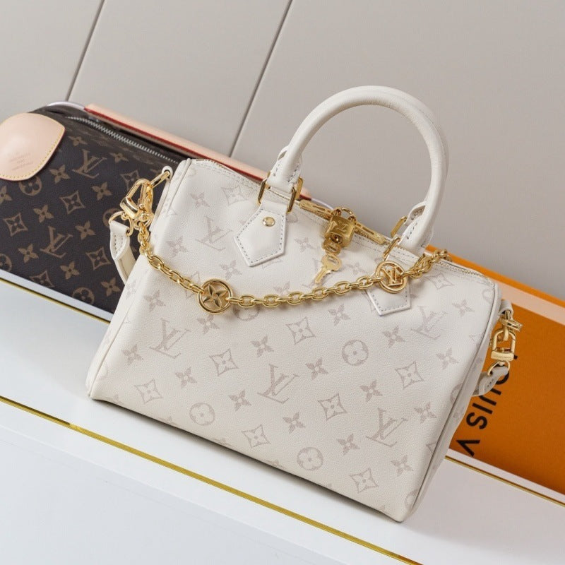 A cream-colored Louis Vuitton monogram Speedy handbag with gold-tone hardware and a chain strap, displayed on a white surface next to a brown monogram duffel bag and an orange book.