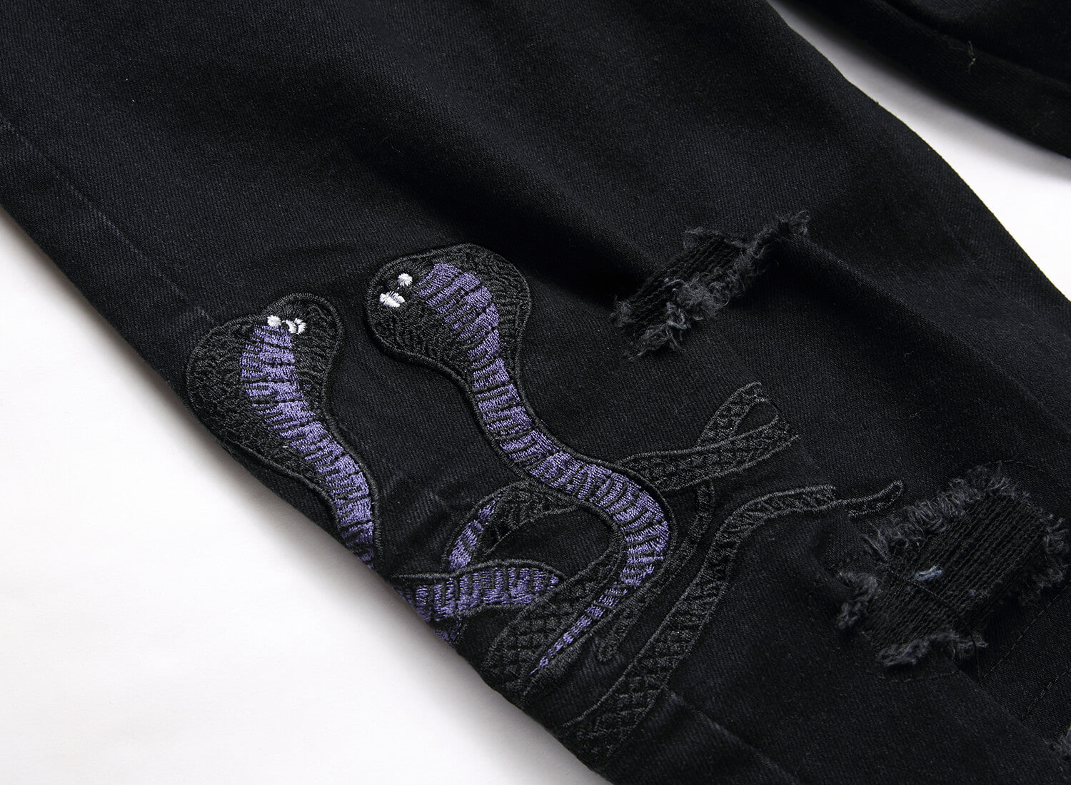 Close-up of black distressed denim featuring a large embroidered purple snake design with small silver bead eyes on the leg.