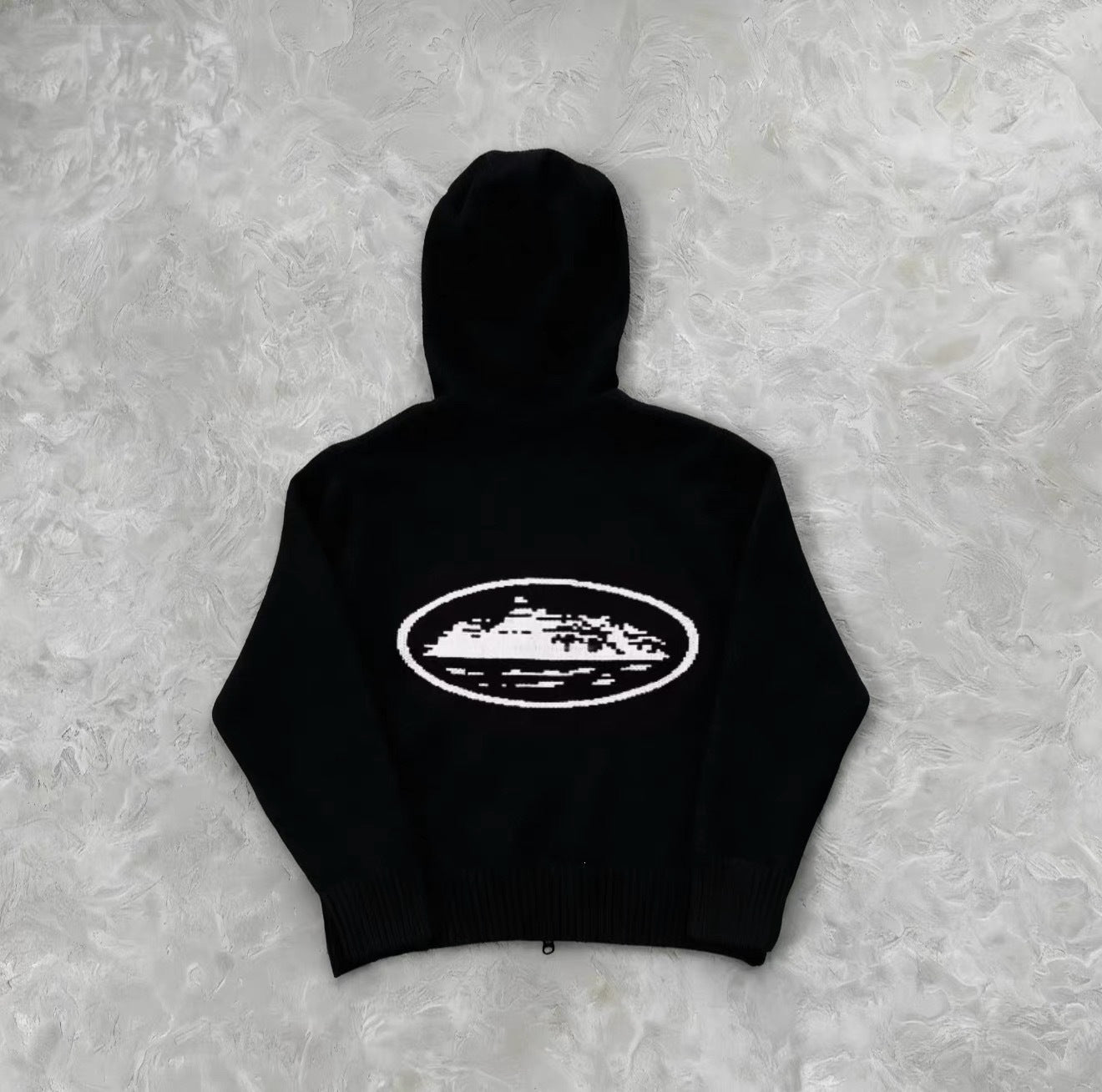 A black knit zip-up hoodie laid flat on a white textured faux fur background, featuring a white pixelated mountain graphic inside an oval on the back.
