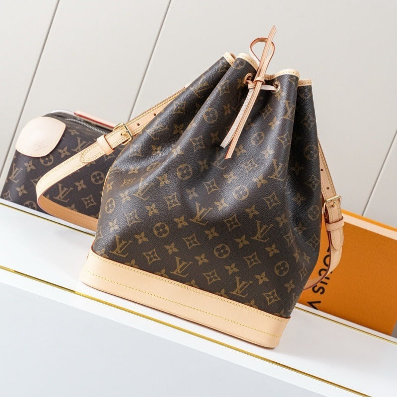 Louis Vuitton monogram canvas bucket bag with beige leather trim and drawstring closure, displayed on a white shelf next to an orange box.