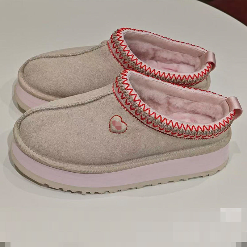 A pair of women's slip-on slippers in light beige suede with pink faux fur lining, decorated with red and gray zigzag trim around the collar and a small pink heart patch on the side, shown on a white surface.