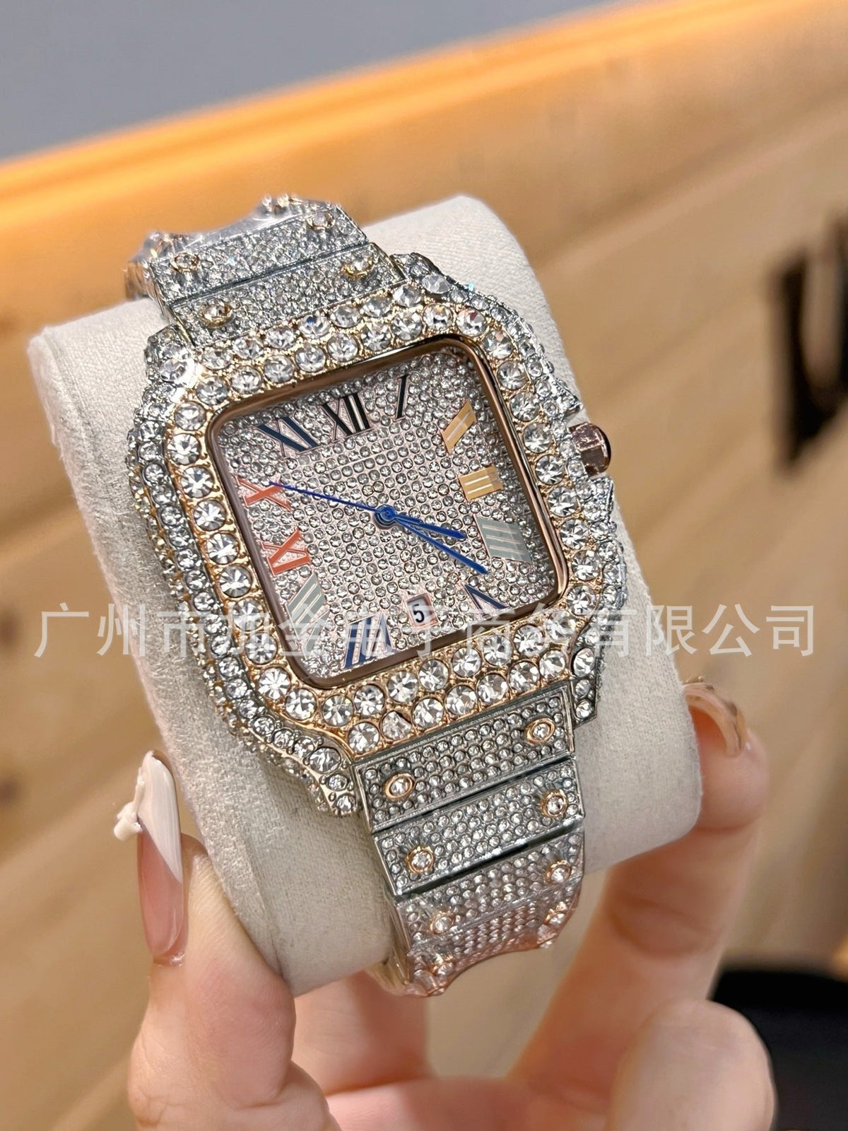 A heavily rhinestone-embellished square analog watch with a silver-toned metal case and matching bracelet, displayed on a light cream wrist strap and held by a hand. The watch face features blue hands and a small colorful dial detail.