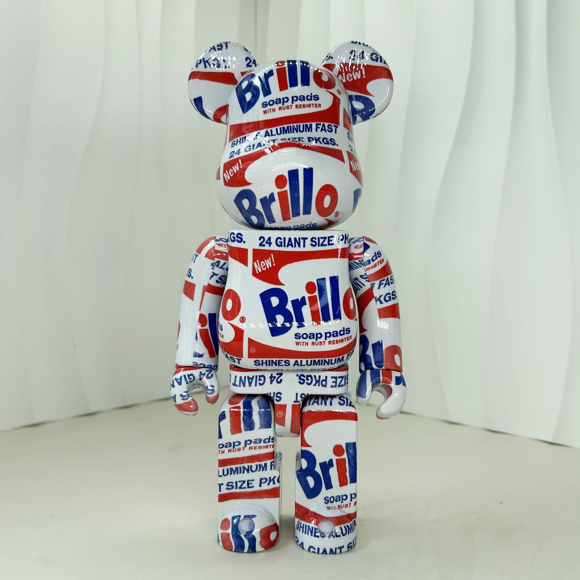 A collectible bear-shaped collectible figure covered in a red, white, and blue Brillo soap dish print pattern, standing against a white curtain backdrop.